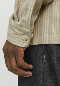 Tessuto a righe beige con una trama testurizzata e lavorazione allentata; presenta due bottoni decorativi rotondi al polsino; mano appoggiata su denim scuro.
