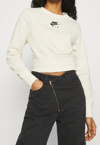 Sweat-shirt court crème avec logo noir "Nike Air", encolure ronde, manches longues et texture lisse, associé à un pantalon sombre taille haute avec une fermeture éclair dorée.