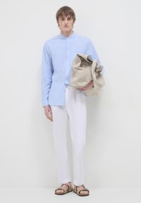 Camicia azzurra a righe con bottoni, pantaloni bianchi e sandali beige. Il modello tiene una borsa di tessuto beige, con una trama morbida e lunghe cinghie.