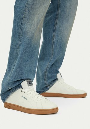 Białe niskie trampki Reebok z gumowymi podeszwami, noszone z luźnymi niebieskimi jeansami, sfotografowane na jednolitym białym tle.