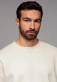 Felpa beige chiaro con scollo a girocollo e una sottile texture. Il modello ha i capelli scuri e una barba corta. Design semplice e pulito.