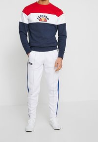 Námořnická mikina s červeno-bílo-černým barevným blokem, která obsahuje logo. Bílé kalhoty s modrými bočními pruhy a černou nášivkou loga.