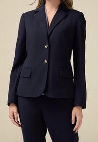 Blazer bleu marine en tissu lisse, doté d'une coupe cintrée, de revers crantés et de deux boutons couleur or. Comprend des détails subtils sur les poches.