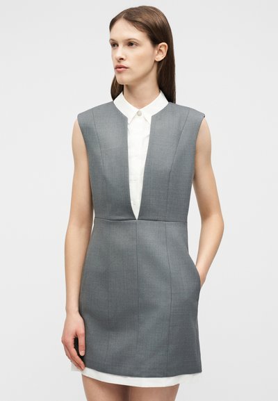 Γυναίκα που φοράει αμάνικο γκρι φόρεμα με βαθύ V λαιμόκοψη πάνω από λευκό πουκάμισο με γιακά, στέκεται με το ένα χέρι στην τσέπη.