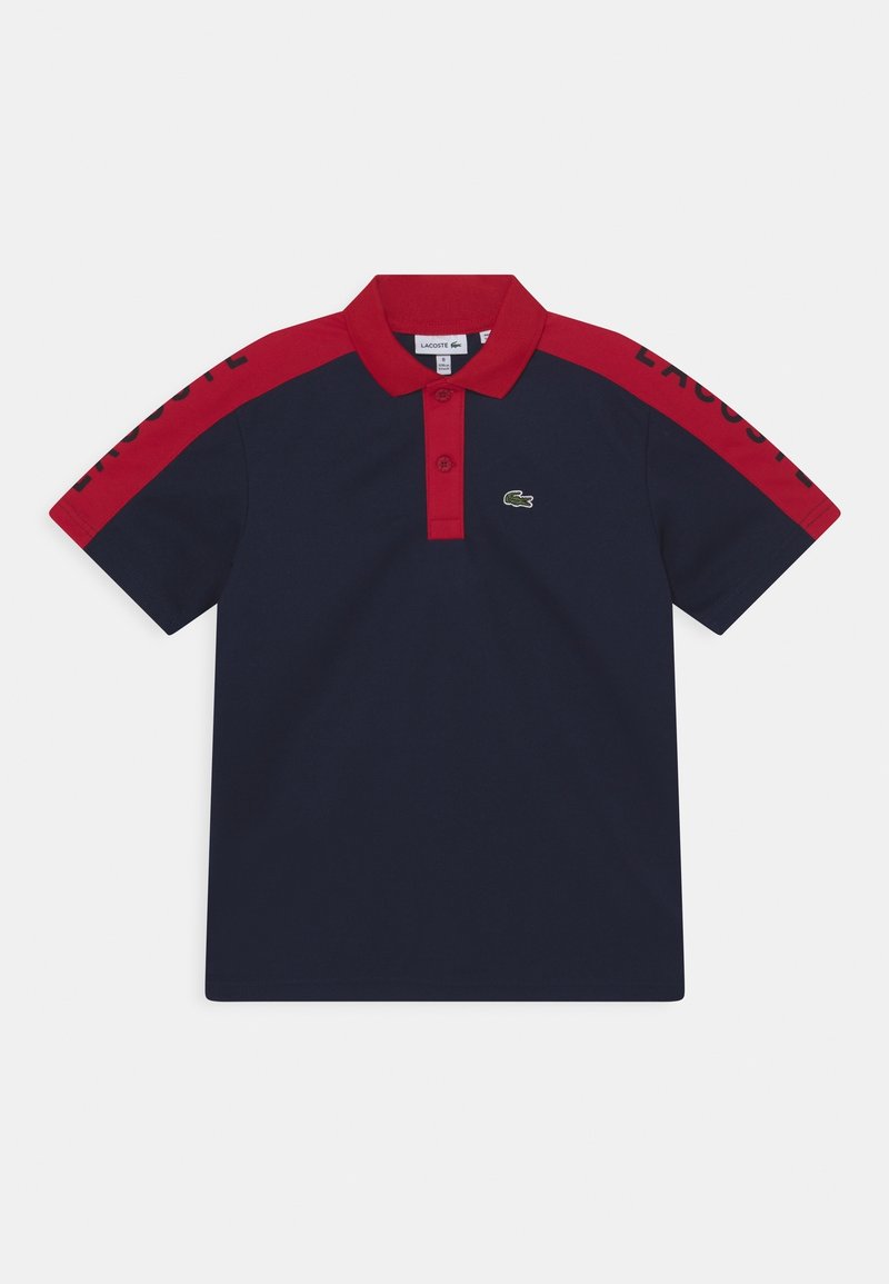 Námořnické modré polo tričko s červeným límcem a pruhy na ramenech, dvoupákový zapínací systém a malým zeleným krokodýlem na hrudi.