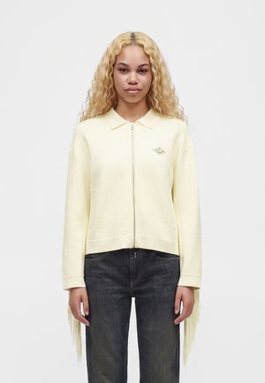Jeune femme aux longs cheveux blonds bouclés, portant un pull zippé jaune pâle avec des franges aux manches et un jean foncé, debout devant un fond blanc.