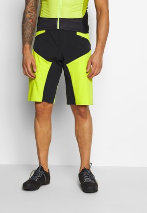Mand iført sorte og skarpt gule cykelshorts med sorte vandresko, stående mod en ensfarvet hvid baggrund.