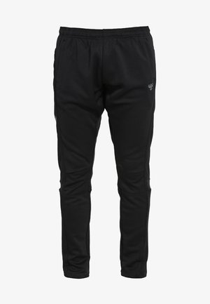 Homme athlétique courant en débardeur noir, pantalon de survêtement noir avec un motif chevron sur le côté, et chaussures de course noires sur un fond uni.