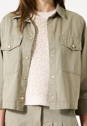 Femme portant un pull en maille beige clair sous une veste kaki boutonnée ouverte avec des poches avant et un pantalon taille haute assorti.