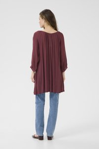 Tunique bordeaux avec dos froncé, manches longues et coupe ample, associée à un jean en denim bleu clair et des chaussures plates sombres.