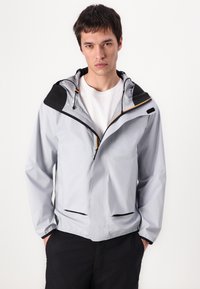 Jachetă waterproof gri deschis, cu glugă neagră și accente negre, cu fermoar în față, două buzunare inferioare și ajustări cu șnur.