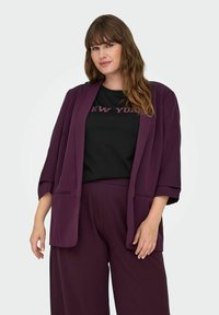 Tamsiai violetinė, laisvai krentanti švarkas su plačiomis rankovėmis ir priekinėmis kišenėmis, dėvima ant juodo marškinėlio su užrašu "NEW YORK" blizgučiais.