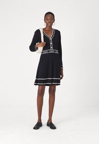 Robe noire à côtes avec manches longues, décolleté en V, boutons dorés, finitions blanches contrastantes, et ceinture avec logo. Associée à des mocassins noirs et un sac beige.