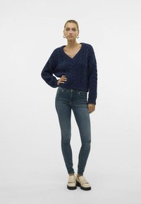 Navy cable-knit sweater with a V-neck design, paired with fitted blue jeans and beige loafers with tassel accents.