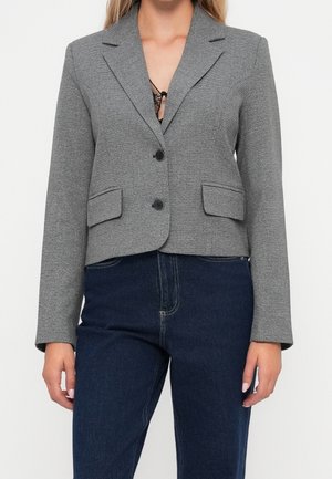 Femme portant un blazer gris à deux boutons sur un top en dentelle, associé à un jean bleu foncé taille haute, debout devant un fond uni.