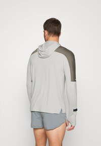 Sudadera con capucha de manga larga en gris claro con detalles en verde oscuro en los hombros y capucha. Cuenta con una abertura lateral y orificios para el pulgar en los puños.
