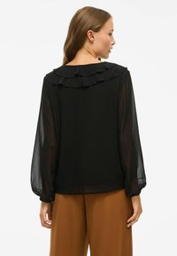 Blusa nera con colletto a strati arricciato, maniche trasparenti e orlo dritto. Abbinata a pantaloni larghi marroni.