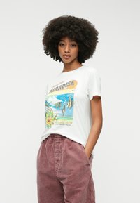 Fehér pamut póló, amely színes grafikai nyomattal van díszítve, trópusi halakkal és virágokkal, puha, rózsaszín kordbársony nadrággal párosítva.