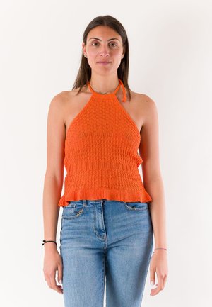 Donna che indossa un top arancione lavorato a maglia abbinato a jeans blu, in piedi su uno sfondo chiaro e uniforme.