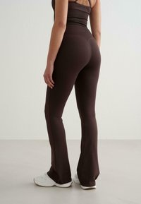 Femme portant un legging ajusté marron foncé et un crop top assorti avec des baskets blanches, se tenant devant un mur clair.