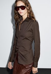 Braunes Hemd mit Knopfleiste in taillierter Passform, langen Ärmeln und glatter Textur. Kombiniert mit einem Lederskirt und übergroßen roten Sonnenbrillen.