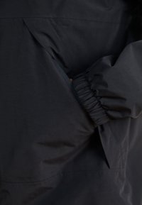 Zwarte jas met elastische manchetten en een opening voor een zijzak, gemaakt van gladde stof met een matte textuur.