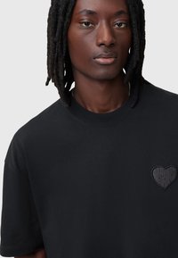 Camiseta de algodón negra con cuello redondo y mangas cortas. Detalle de logo en forma de corazón en relieve en la parte izquierda del pecho. Textura suave.