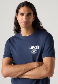 Tumši zils kokvilnas T-krekls ar baltu "LEVI'S®" logo un apļveida grafiku uz krūšu daļas, �īsām piedurknēm un apaļu kakla izgriezumu.