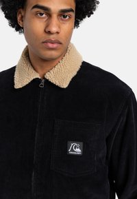 Sort fløjlsjakke med en beige sherpa-krave, lynlåslukning og en brystlomme med et logo-lap. Tekstiler med ribbet design.