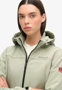 Lichtgroene softshell jack met een ritssluiting aan de voorkant, verstelbare capuchon en logoprints. Stikdetails voegen textuur en structuur toe.