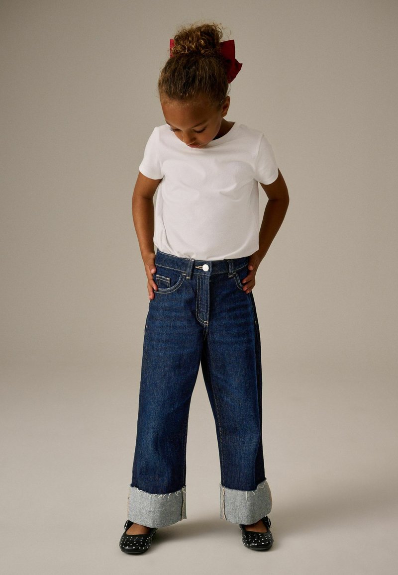 Jeans in denim con vestibilità ampia, colore blu scuro, orli sfilacciati e dettagli in chambray azzurro chiaro ai polsi, abbinati a una t-shirt bianca semplice.