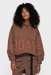 Hoodie marron avec un grand texte "10DAYS" dans une teinte contrastante. Doté d'une capuche avec cordon et d'une coupe décontractée. Porté avec un short assorti.