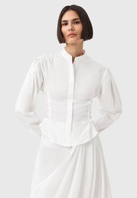 Camicia bianca con colletto alto, maniche a sbuffo e dettagli arricciati ai lati. Realizzata in morbido cotone, con vestibilità sartoriale.