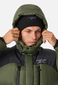 Green puffer jacket with a black patterned panel, featuring a high collar, adjustable hood, and zippered pockets. Black knit beanie included.