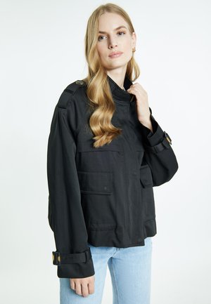 Femme blonde aux cheveux ondulés portant une veste noire avec de grandes poches et des poignets bouclés, associée à un jean bleu clair, sur fond blanc.