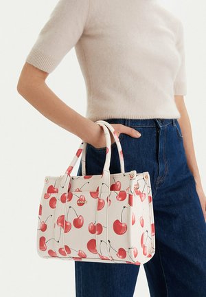 Kobieta trzymająca białą torebkę z czerwonym nadrukiem wisienek, ubrana w kremowy sweter z krótkim rękawem i niebieskie dżinsy, przycięte na wysokości talii.