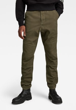 Pantalon vert olive en tissu texturé, avec des renforts aux genoux, des poches latérales et une coupe décontractée, porté avec des bottes noires.