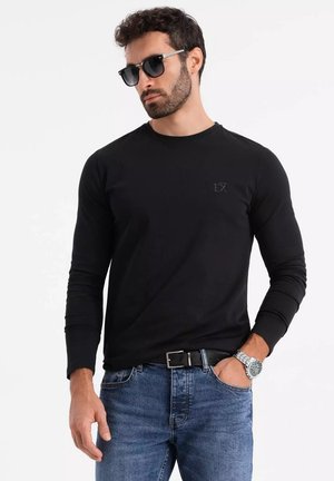 Schwarzes Langarmshirt mit ribbed Rundhalsausschnitt und dezentem Logo auf der Brust, kombiniert mit blauen Jeans und schwarzen Sonnenbrillen.