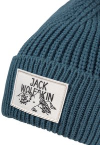 Teal gebreide beanie met ribbelstructuur, voorzien van een witte patch met het label "JACK WOLFSKIN" en een bergafbeelding in zwarte borduursel.