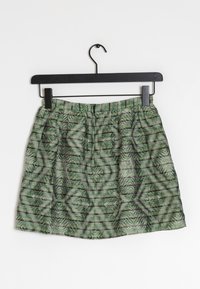 Grön och svart geometriskt mönstrad kjol med elastisk midja hängande på en svart galge mot en vit bakgrund.