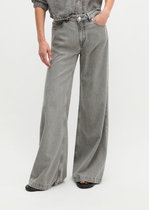 Persoon draagt grijze denim jeans met hoge taille en wijde pijpen, één hand in de zak en zichtbare zwarte sandalen met open teen.