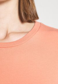 Gros plan sur le cou et l'épaule d'une personne portant un haut col rond couleur corail clair avec des cheveux bruns raides jusqu'aux épaules.