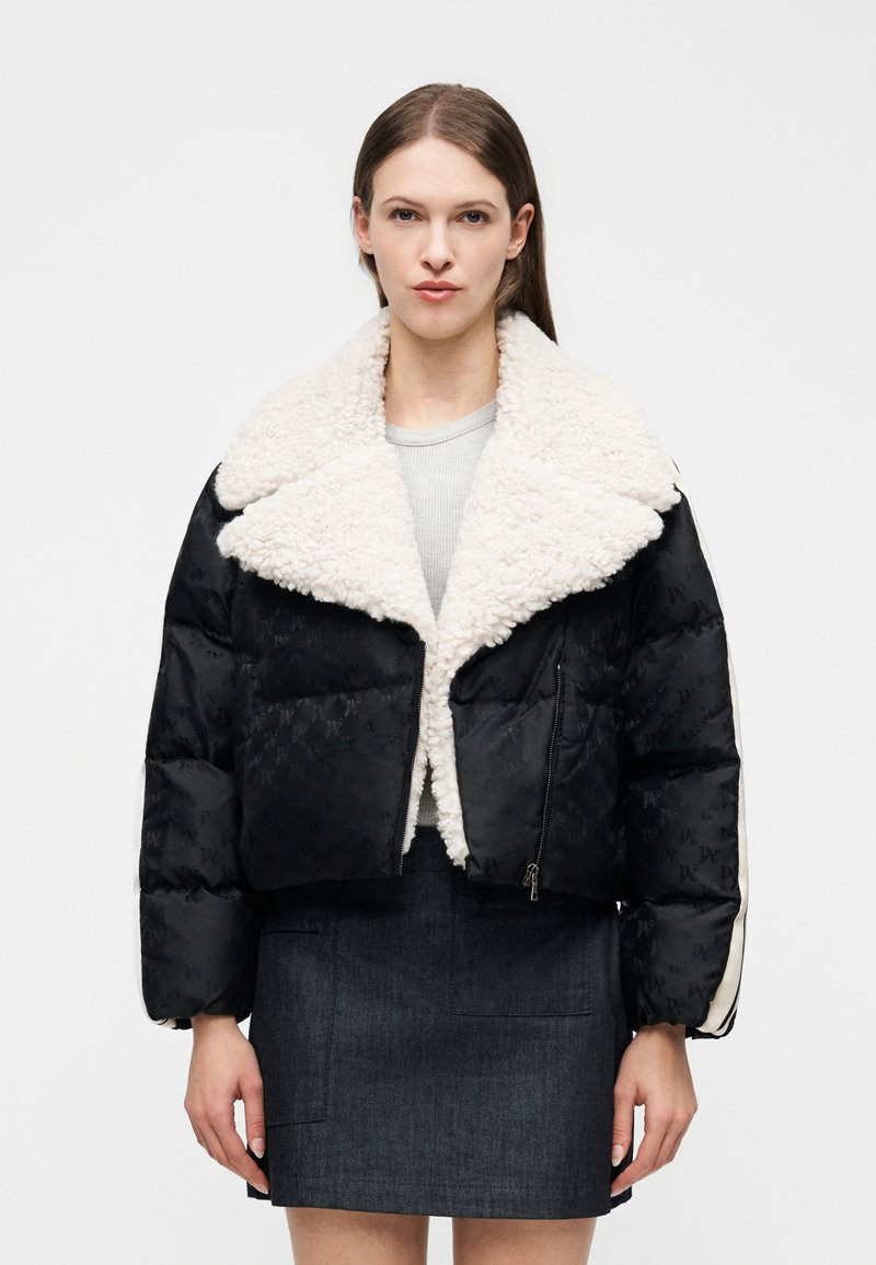 Γυναίκα που φοράει μια μαύρη φουσκ ωτή ζακέτα με μεγάλο λευκό γιακά από συνθετική γούνα, ανοιχτό γκρι μπλούζα και σκουρόχρωμη τζιν φούστα, στραμμένη προς τα εμπρός πάνω σε λευκό φόντο.