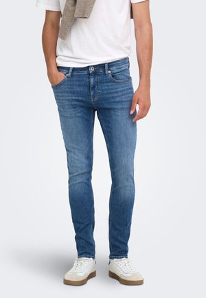 Homme portant un jean slim bleu, des baskets blanches et un t-shirt blanc avec un pull beige posé sur une épaule.