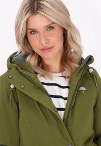 Olivgrön jacka med huva, grå foder och silverfärgade tryckknappar. Har en liten vit paraplylogotyp på bröstet och dragkedjestängning.