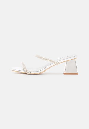 White heeled sandals with a glossy finish, featuring two thin straps adorned with rhinestones and a metallic triangular block heel.