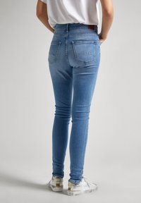 Los jeans ajustados de color azul claro tienen una cintura alta, bolsillos traseros y detalles de costura sutiles. Se combinan con zapatillas blancas y una camisa blanca holgada.