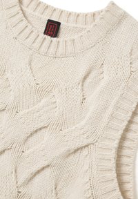 Cremfarbener, dicke Kabelstrickpullover mit geripptem Rundhalsausschnitt und rotem sowie schwarzem Markenetikett im Kragen.