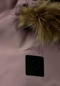 Ljust rosa tyg med en mjuk textur, med en brun pälsbesättning och ett svart, sköldformad logotyp-patch i nedre hörnet.