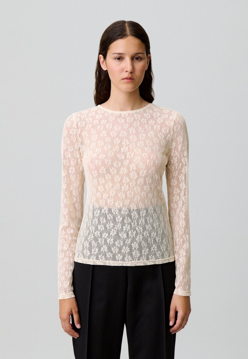 Mujer con pelo oscuro y lacio que lleva una blusa de encaje floral crema transparente de manga larga sobre un sujetador, combinada con pantalones negros de cintura alta, contra un fondo sencillo.